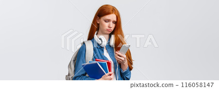 Silly sad and gloomy pouting girl with red foxy hair, frowning sighing and looking with regret smartphone screen, reading troublesome message, feeling bad, stand with backpack and notebooks 116058147