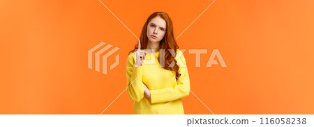 Redhead girl express disapproval as shaking her finger at someone with disappointed angry face, frowning give warning, prohibit unacceptable behaviour, standing displeased orange background 116058238