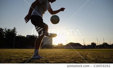 Professional footballer juggling soccer ball on stadium at sunset. Young man kicking ball at green field. Sportsman practicing tricks at meadow with sunlight at background. Freestyle football concept 116058262