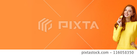 Waist-up portrait lovely redhead girl holding tasty yellow macaron near mouth and smiling joyfully, like eating sweets, buy dessert for romantic valentines date, posing cheerful orange background 116058350