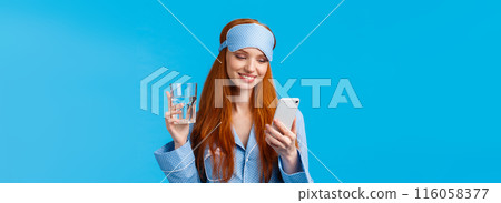 Happy and relaxed attractive feminine redhead woman holding smartphone and glass water, smiling joyfully and delighted as reading nice lovely message from mobile application, blue background Happy and relaxed attractive feminine redhead woman holding smartphone and glass water, smiling joyfully and delighted as reading nice lovely message from mobile application, blue background 116058377