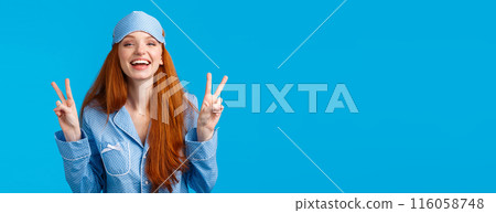 Happy and carefree feminine young redhead female in nightwear, sleep mask and pyjama, laughing joyfully, feeling self-assured and pretty showing peace signs, smiling toothy, blue background 116058748