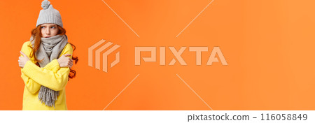 Its freezing cold outside. Cute redhead girl shaking and embracing herself to warm up, waiting someone on street in grey scarf, winter hat during snow and low temprature weather, orange background 116058849
