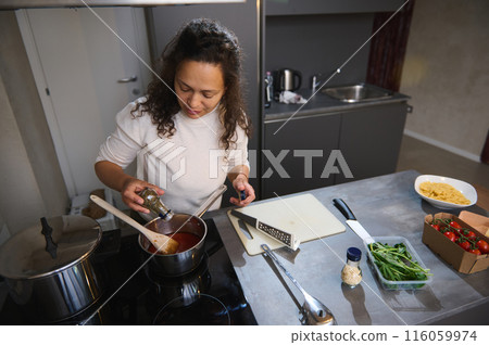 View from above of happy housewife adding culinary herbs, condiments, seasoning the tomato sauce in a saucepan, standing at electric kitchen stove in home interior. Food preparation concept View from above of happy housewife adding culinary herbs, condiments, seasoning the tomato sauce in a saucepan, standing at electric kitchen stove in home interior. Food preparation concept 116059974
