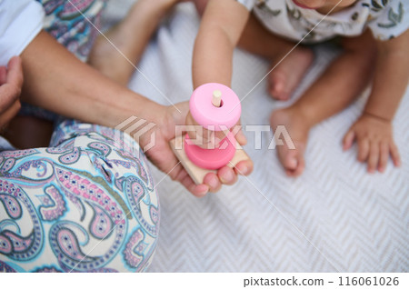 Focus on wooden pyramid in woman's hands, playing with her child educational games, stacking round circles, sitting together on linen blanket outdoors. Eco toys for kids development and entertainment Focus on wooden pyramid in woman's hands, playing with her child educational games, stacking round circles, sitting together on linen blanket outdoors. Eco toys for kids development and entertainment 116061026