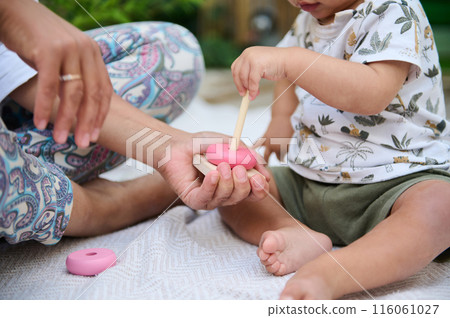Close-up with focus on hands of a baby boy stacking round circles of educational developmental wooden pyramid, playing with his mother supervising him, sitting together on the linen blanket outdoors 116061027
