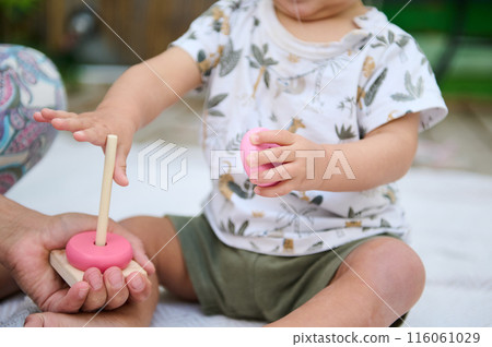 Close-up with focus on hands of a toddler stacking round circles of educational developmental wooden pyramid, playing with his mother supervising him, sitting together on the linen blanket outdoors Close-up with focus on hands of a toddler stacking round circles of educational developmental wooden pyramid, playing with his mother supervising him, sitting together on the linen blanket outdoors 116061029