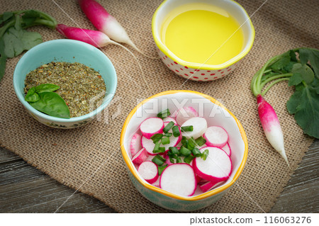 Salad of fresh garden grown radishes (French breakfast variety) and green onion in a little bowl on the wooden table with burlap cloth, ingredients with oil and dry herbs around. Salad of fresh garden grown radishes (French breakfast variety) and green onion in a little bowl on the wooden table with burlap cloth, ingredients with oil and dry herbs around. 116063276