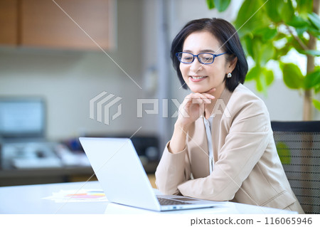 A middle-aged woman wearing reading glasses looking at a computer in the office A middle-aged woman wearing reading glasses looking at a computer in the office 116065946