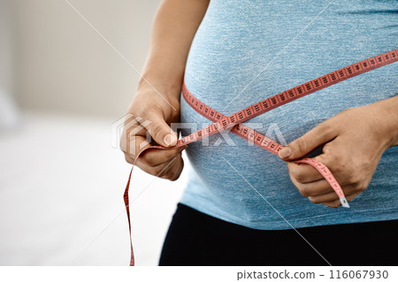 Close-up of pregnant woman in blue tank top measuring her belly with pink tape measure, emphasizing growth and prenatal care Close-up of pregnant woman in blue tank top measuring her belly with pink tape measure, emphasizing growth and prenatal care 116067930