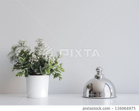 A white potted plant with green leaves and white flowers sits next to a shiny silver bell on a white surface. 116068975