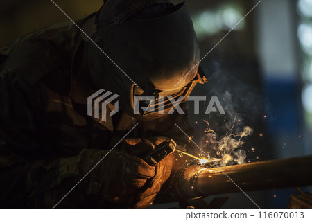 Welder at work. Man in a protective mask. The welder makes seams on the metal. Sparks and smoke when welding. Welder at work. Man in a protective mask. The welder makes seams on the metal. Sparks and smoke when welding. 116070013