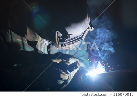 Welder at work. Man in a protective mask. The welder makes seams on the metal. Sparks and smoke when welding. Welder at work. Man in a protective mask. The welder makes seams on the metal. Sparks and smoke when welding. 116070015