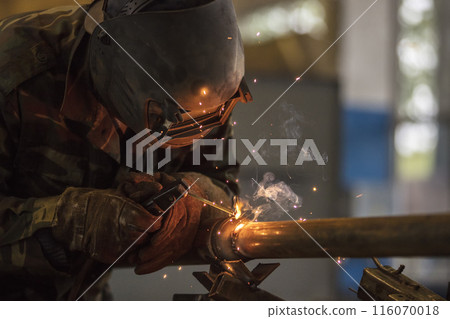 Welder at work. Man in a protective mask. The welder makes seams on the metal. Sparks and smoke when welding. Welder at work. Man in a protective mask. The welder makes seams on the metal. Sparks and smoke when welding. 116070018