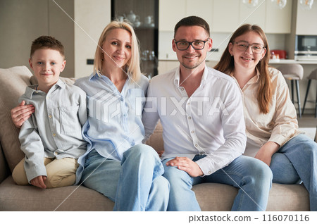 Big family chilling together on cream sofa. Cute family portrait of four members looking to the camera. Happy moments of close people in modern living room. 116070116