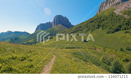 Natural mountain scenery. The trail passes through green grass. A majestic cliff rises ahead. 116071215