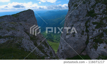 Mountain peaks rise majestically beneath a clear blue sky, and a serene green valley spreads out below. View across the cliffs. Mountain peaks rise majestically beneath a clear blue sky, and a serene green valley spreads out below. View across the cliffs. 116071217