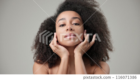 Beauty and healthcare concept - beautiful African American woman with curly afro hairstyle and clean, healthy skin touching face with hand, doing facial massage, posing and looking at camera Beauty and healthcare concept - beautiful African American woman with curly afro hairstyle and clean, healthy skin touching face with hand, doing facial massage, posing and looking at camera 116071386