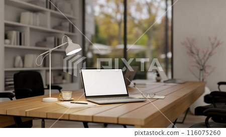 A laptop computer with a white-screen mockup on a wooden meeting table in a modern meeting room. 116075260