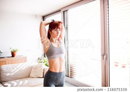 Beautiful redhead woman's home fitness routine. Woman stretching after workout, wearing sportswear. 116075339