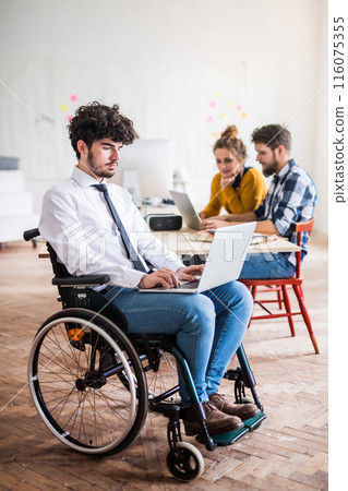 Young man in wheelchair working with colleagues on a new project in a startup company office. Working in co-working space. Young man in wheelchair working with colleagues on a new project in a startup company office. Working in co-working space. 116075355