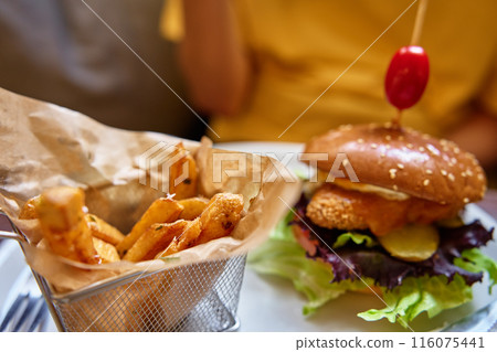 Close-up of tasty chicken burger with side of crispy fries in basket served on white plate in restaurant. Fast food and junk food concept. 116075441