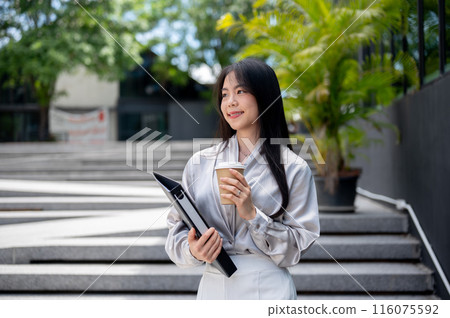 A happy Asian businesswoman with a file folder and a takeaway coffee cup is walking down the stairs. 116075592