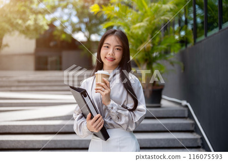 Attractive Asian businesswoman with a file folder and a coffee cup, standing on the stairs outdoors. Attractive Asian businesswoman with a file folder and a coffee cup, standing on the stairs outdoors. 116075593