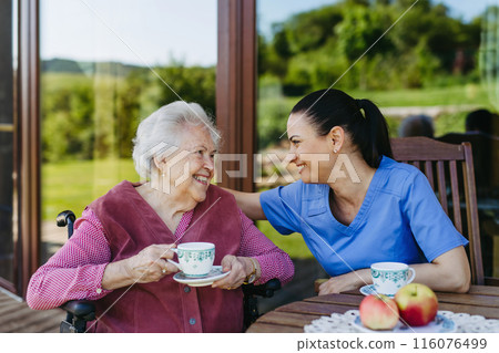 Female caregiver spending time with elderly woman, drinking coffee and talking. Nurse and senior woman in wheelchair enjoying a warm day outdoors. Female caregiver spending time with elderly woman, drinking coffee and talking. Nurse and senior woman in wheelchair enjoying a warm day outdoors. 116076499