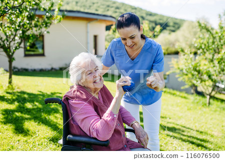 Female caregiver doing motorized exercises with senior woman in wheelchair. Nurse and elderly woman squeezing small, soft ball. 116076500