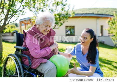 Female caregiver doing motorized exercises with senior woman in wheelchair. Nurse and elderly woman working out with exercise ball. 116076525