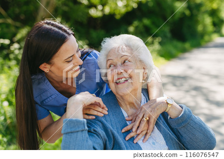 Female caregiver pushing senior woman in wheelchair. Nurse and elderly woman enjoying a warm day in nursing home, public park. 116076547