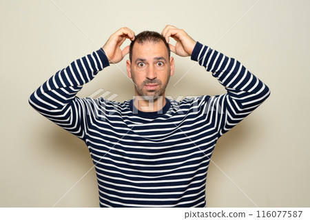 Bearded Hispanic man in his 40s wearing a striped sweater with a serious face and hands on his head worried about an adverse situation, isolated over beige background. 116077587