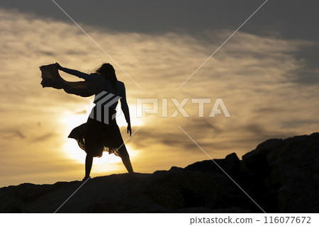 A woman is standing on a rocky hillside, with the sun setting in the background A woman is standing on a rocky hillside, with the sun setting in the background 116077672