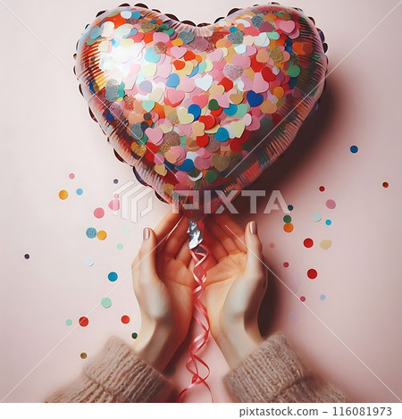 A heart-shaped balloon in female hands on a pink surface surrounded by scattered confetti. Joyful and festive atmosphere 116081973