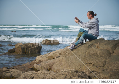 Man sitting on rocky shoreline taking photo with smartphone at the beach Man sitting on rocky shoreline taking photo with smartphone at the beach 116082337