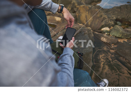 Person using smartphone outdoors while sitting on rocky terrain wearing casual attire Person using smartphone outdoors while sitting on rocky terrain wearing casual attire 116082372