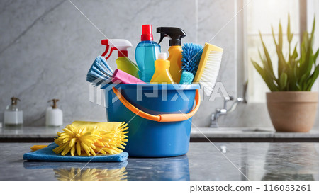 A cleaning caddy filled with various cleaning supplies, including spray bottles, brushes, and cloths, set on a marble countertop in a brightly lit kitchen. 116083261