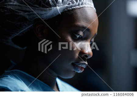 Side view closeup of young Black woman wearing protective clothing in operating room lit by blue light copy space 116084006