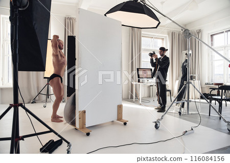 Wide angle view at male bodybuilder posing for camera during professional photoshoot with photographer taking pictures in minimal white studio, copy space Wide angle view at male bodybuilder posing for camera during professional photoshoot with photographer taking pictures in minimal white studio, copy space 116084156
