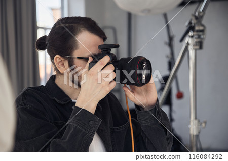 Side view portrait of young male photographer looking into camera viewfinder and taking pictures during photoshoot in minimal studio, copy space 116084292