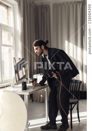 Vertical full length portrait of young professional photographer holding camera and using computer in minimal photo studio with light accents 116084309
