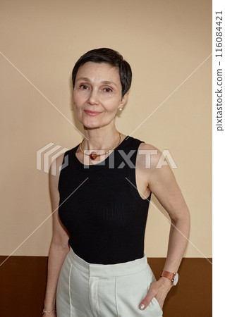 Vertical waist up portrait of elegant mature woman smiling at camera posing against wall in neutral colors shot with flash 116084421