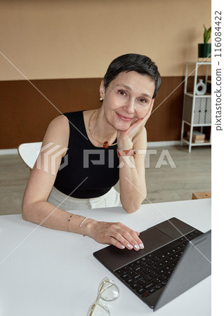 Vertical portrait of successful mature businesswoman smiling at camera while using laptop at desk in office shot with flash 116084422