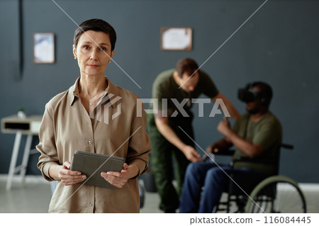 Waist up portrait of mature Caucasian woman as mental health expert looking at camera holding tablet with army veterans in background copy space 116084445