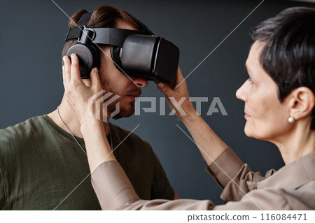 Minimal side view closeup of army veteran wearing VR headset in therapy session with female psychotherapist setting up tech 116084471
