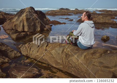 Man sitting on rock by the ocean, enjoying a peaceful moment during a hiking trip 116085232