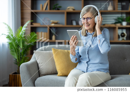Elderly woman enjoying technology, listening to music or a podcast with headphones and using a tablet. She is sitting comfortably on a sofa in a contemporary living room setting. 116085955