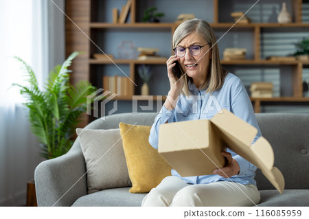 Elderly woman sitting on a couch, talking on a mobile phone, and opening a package. She looks delighted and curious about the contents of the box. Cozy and modern living room setting. Elderly woman sitting on a couch, talking on a mobile phone, and opening a package. She looks delighted and curious about the contents of the box. Cozy and modern living room setting. 116085959