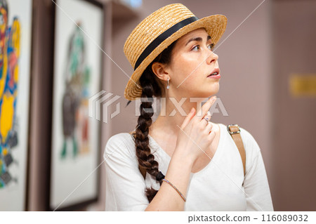 Portrait of young dreamy Caucasian woman in straw hat admiring paintings in the gallery. Concept of cultural education at museum Portrait of young dreamy Caucasian woman in straw hat admiring paintings in the gallery. Concept of cultural education at museum 116089032